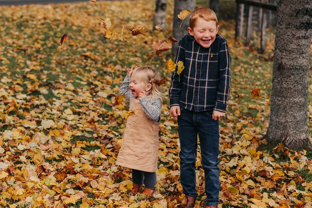 A brother and sister giggling as autumn leaves fall around them